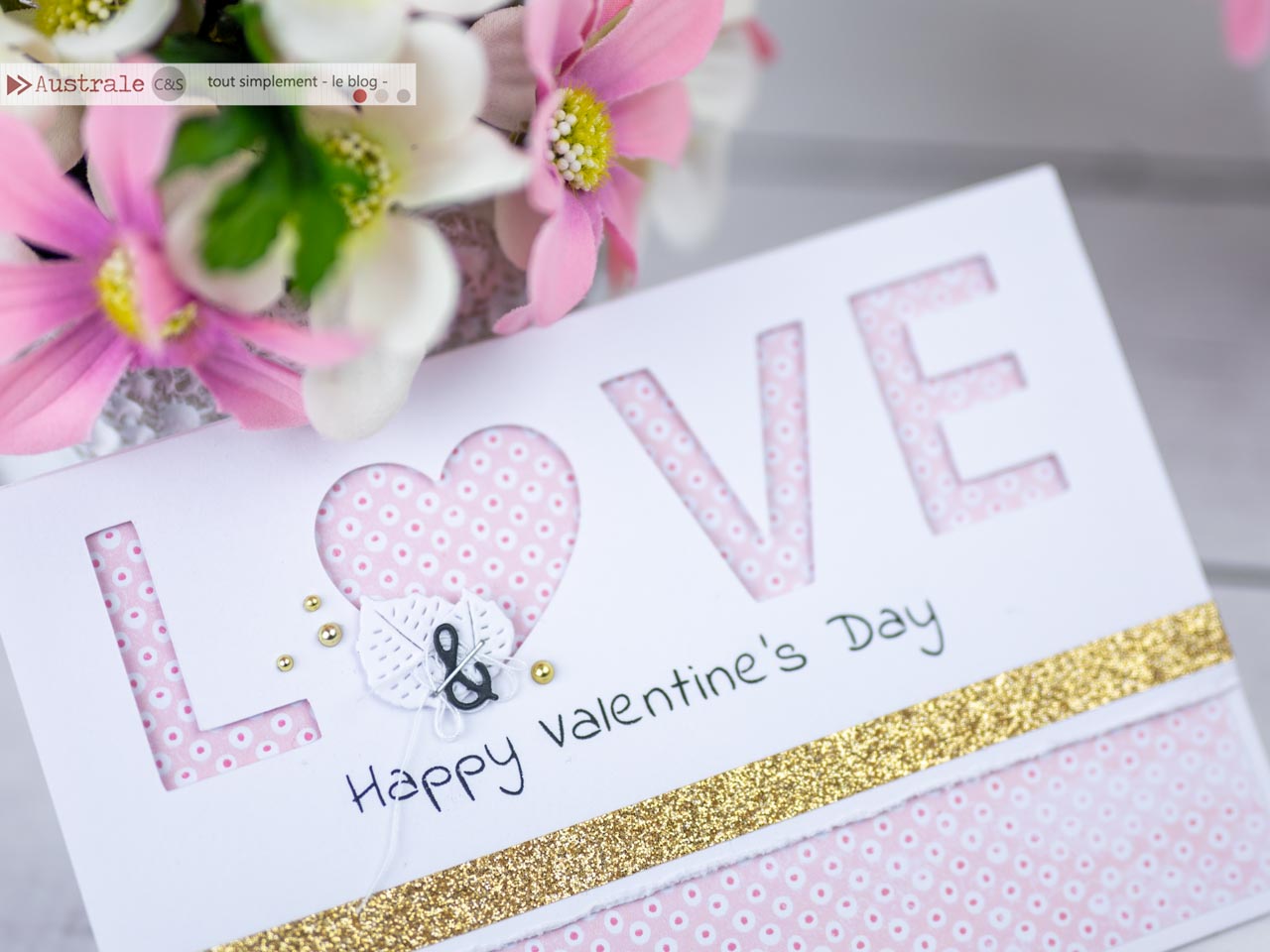 Création d'une carte de Saint-Valentin avec le mot LOVE en transparence sur un papier imprimé de petits ronds roses, le texte happy valentine's day imprimé, une bordure vieillie avec une longueur de masking tape dorée et quelques feuillages, esperluette, fil et agrafe.